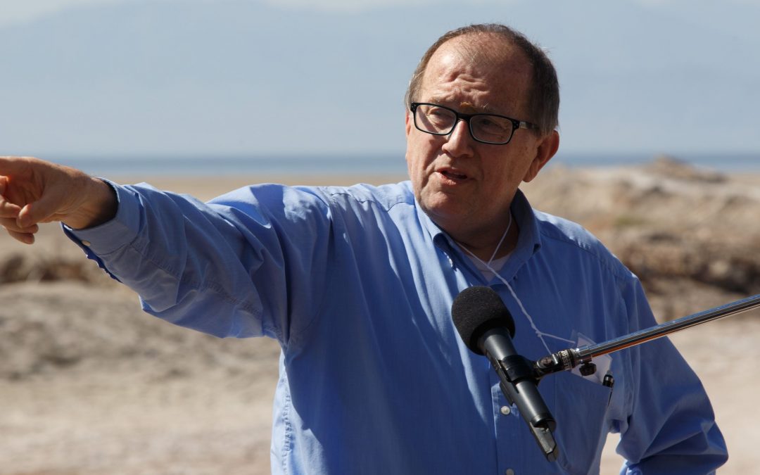 Older man in light blue shirt and wearing glasses speaks into a microphone and points into the distance