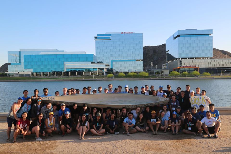 large group of students smiling and posing on the shore of a body of water holding a concrete canoe and a sign that says "UCLA go bruins"